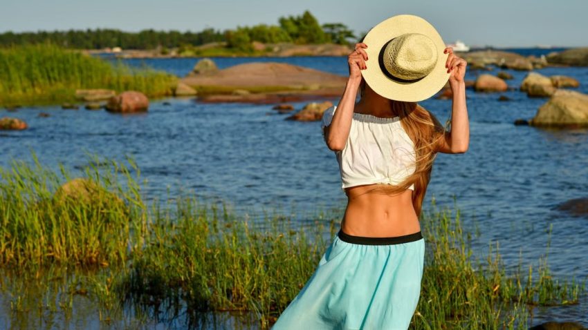 Woman enjoys a summer day by the rocky beach in Helsinki, hiding face with a straw hat